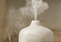 A white ceramic diffuser emits gentle wisps of steam, set against a neutral, blurred background, creating a serene and calming atmosphere.